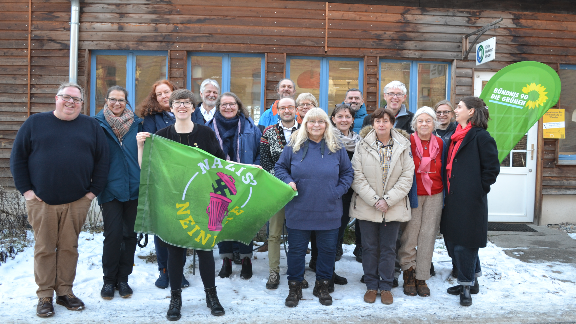 Auf dem Bild sind Teilnehmer:innen der Aufstellungsversammlung der Grünen Werder zu sehen. Insgesamt 17 Personen stehen vor der Klimawerkstatt Werder. Vorne auf dem Bild hält eine Frau ein Banner mit der Aufschrift "Nazis? - Nein danke!"